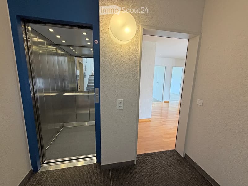 Hallway with a modern elevator on the left and an open doorway leading to a room with wooden flooring on the right. The hallway features a round wall-mounted light fixture and a power outlet.