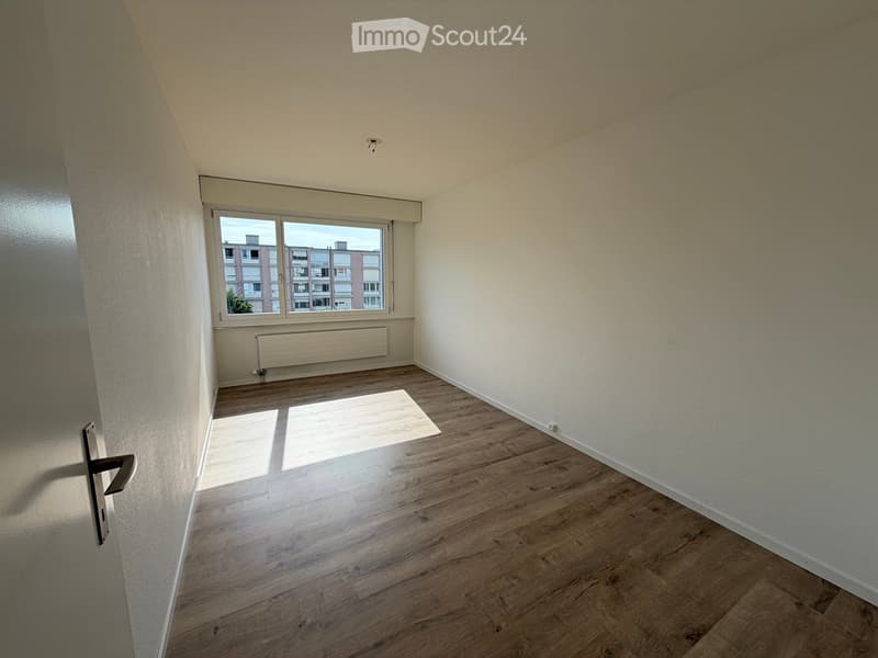 Empty room with wooden flooring, white walls, a large window with a view of buildings, and a radiator below the window.