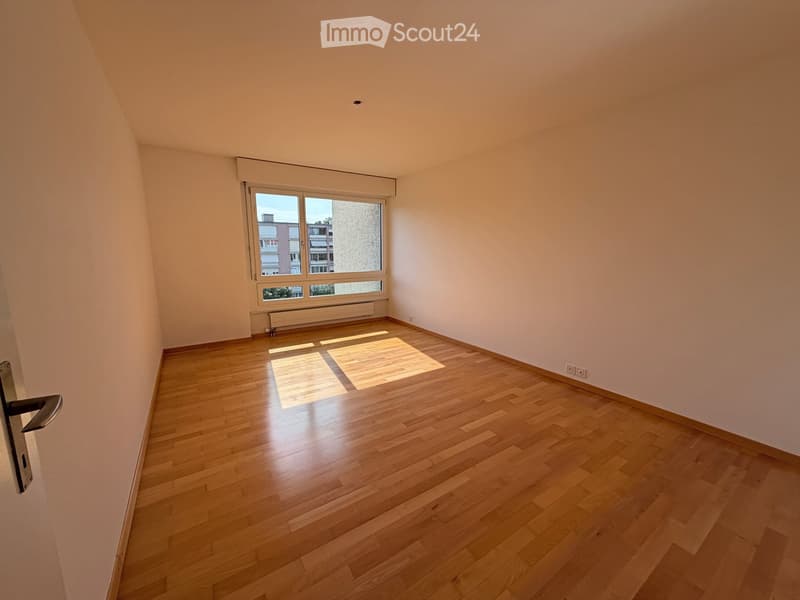Empty room with wooden flooring, white walls, and a large window letting in natural light.