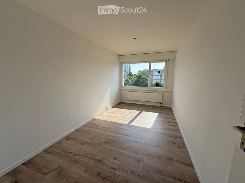 Empty room with wooden flooring, white walls, and a large window offering a view of trees and buildings.