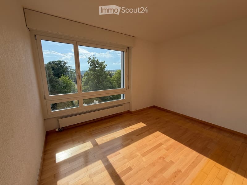 Empty room with wooden flooring and large windows offering a view of trees and sky. No furniture or equipment present.