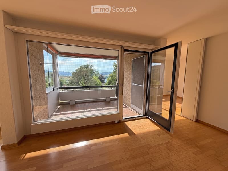 Empty room with wooden flooring, large sliding glass doors leading to a balcony with a scenic view of trees and mountains.
