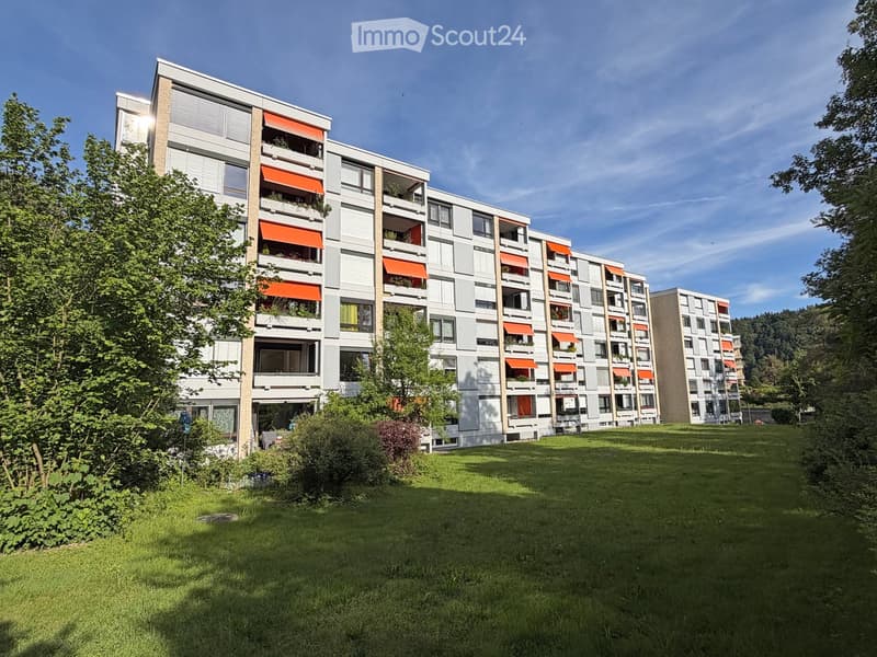 Modern multi-story residential building with balconies, orange awnings, and surrounding greenery.
