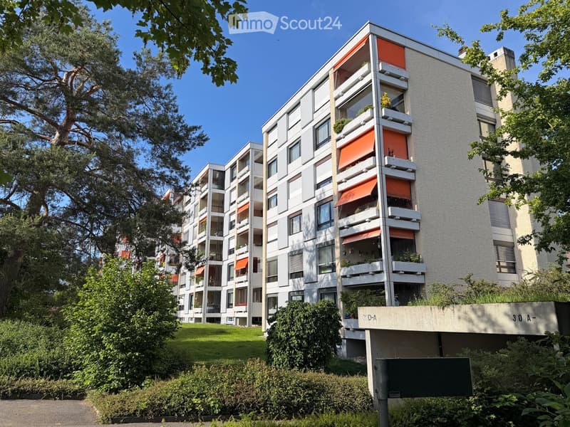 Modern multi-story residential building with balconies, greenery, and trees in the foreground.