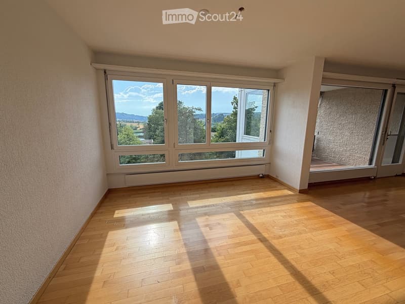 Empty room with wooden flooring, large windows offering a scenic view, and a sliding glass door leading to a balcony area.