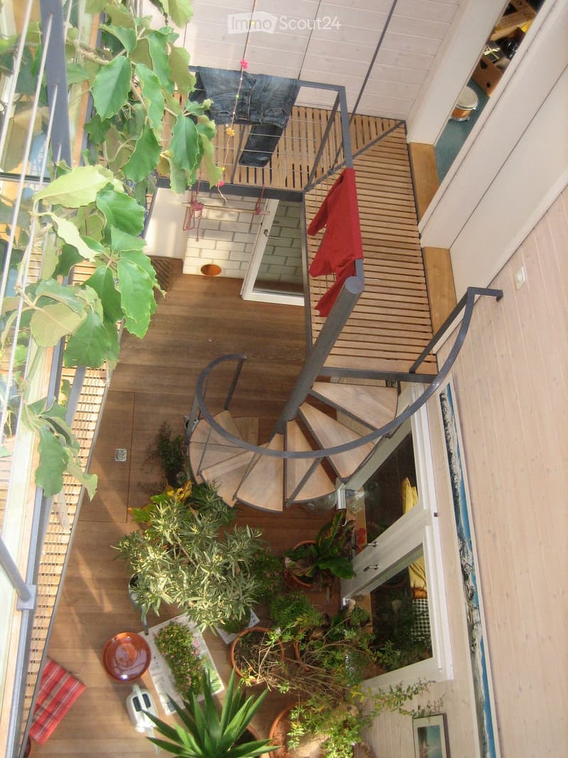 Balcony with wooden decking, metal railing, and multiple potted plants, including leafy greens and herbs. Laundry is hanging on a clothesline, and there is a small table with a bowl and a striped cloth.