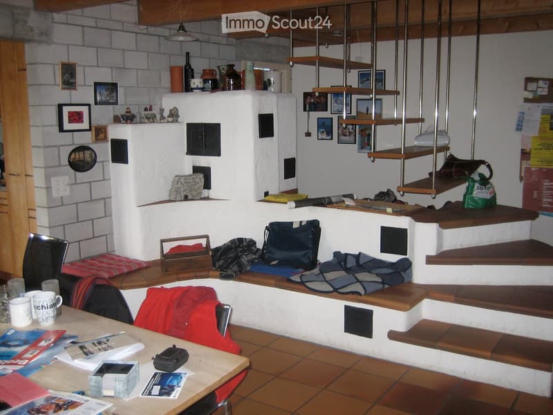 A cozy, rustic living space with a wooden staircase, a built-in bench, and a small table with cups and magazines. The room features exposed brick walls, wooden beams, and various decorative items on shelves.