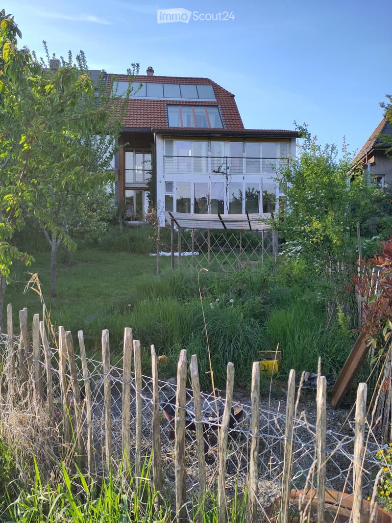 A modern two-story house with large windows and a balcony is surrounded by a lush garden with tall grasses, trees, and a wooden fence.