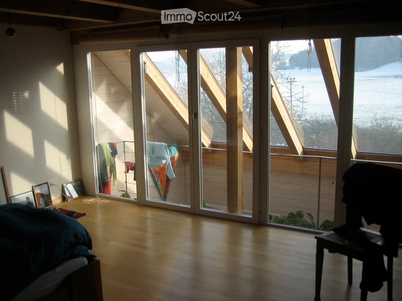 Bright, airy bedroom with wooden floors, large glass sliding doors leading to a balcony, a bed with blue bedding, a small table, a clothing rack, and several picture frames on the wall.