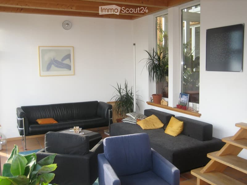Modern living room with a black leather sofa, grey sectional sofa, coffee table, and a small wooden table. The room features large windows with plants visible outside, a potted plant on the floor, and artwork on the walls.