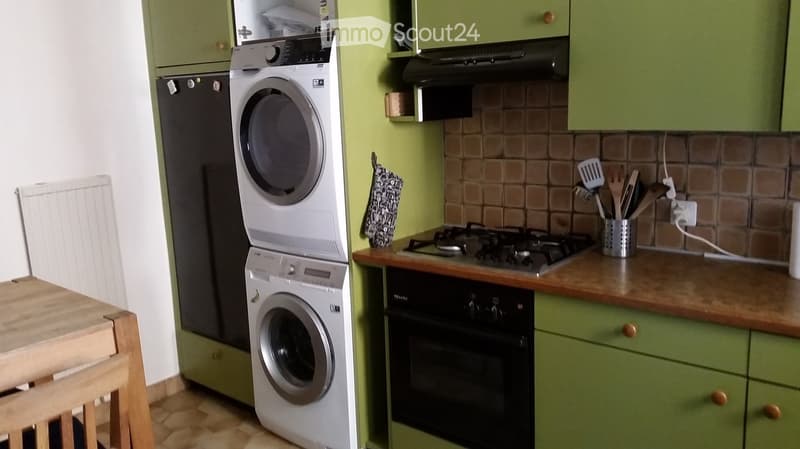 Green kitchen with gas stove, oven, cabinets, washing machine, and a small wooden table with a stool.