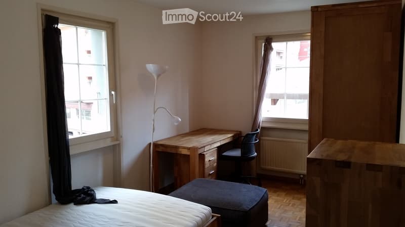 A small, furnished bedroom with a bed, desk, chair, and floor lamp. The room has wooden flooring and two windows with curtains.