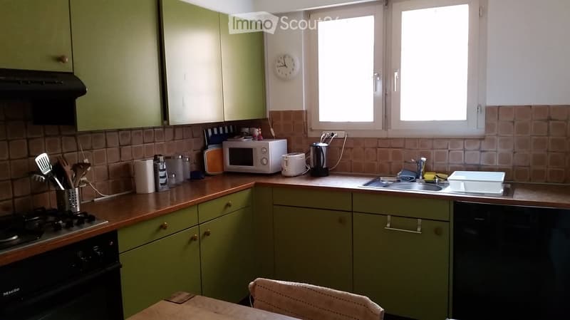 A well-lit kitchen with green and white cabinets, a wooden countertop, and various appliances including an oven, microwave, toaster, and sink. There is a clock on the wall and a stool near the counter.