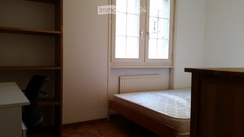 A small bedroom with a single bed, a chair, a radiator, and a shelf. The room has wooden flooring and a window with multiple panes.