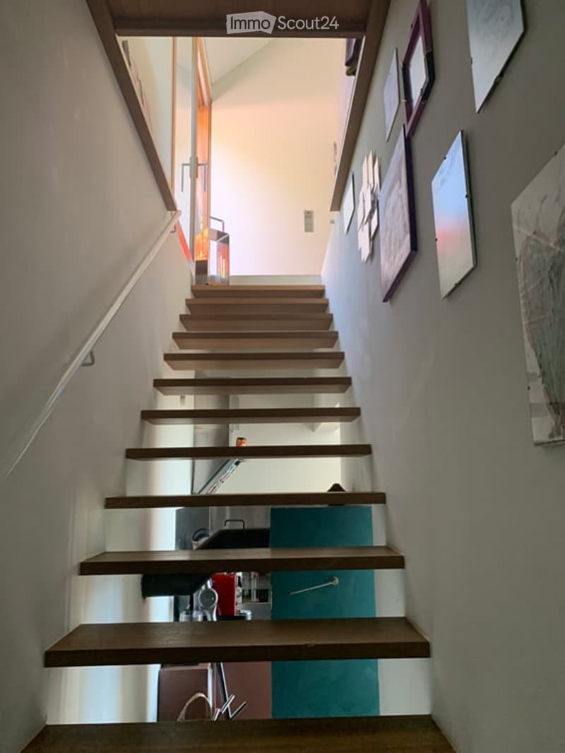 Modern wooden staircase with a sleek design, white walls, and framed artwork on the wall. The staircase features a continuous wooden railing and a clean, minimalist aesthetic.