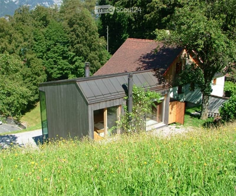 Modern, single-story house with a dark grey exterior and a large glass wall, surrounded by lush greenery and a grassy field.