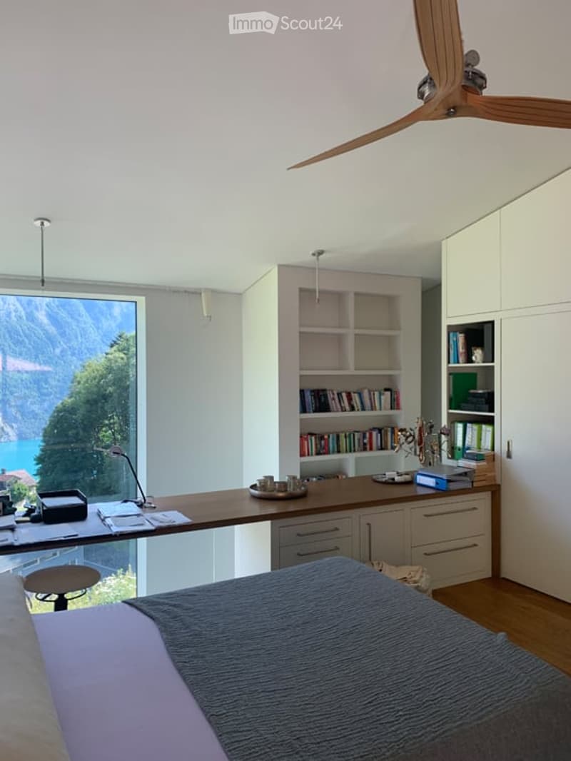 Modern bedroom with a large window offering a scenic mountain view, a bed with a grey duvet, a wooden desk with a chair, a bookshelf filled with books, and a ceiling fan.