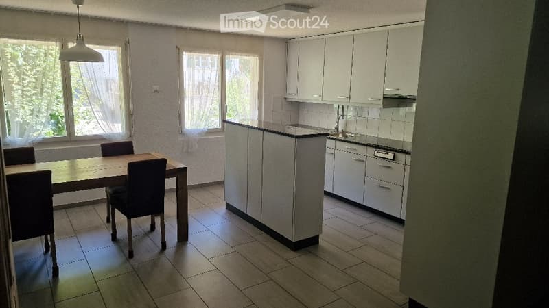 A modern, open-plan kitchen and dining area with white cabinets, a central island, and a dining table with four chairs. The space features large windows with white curtains, tiled flooring, and a hanging pendant light.