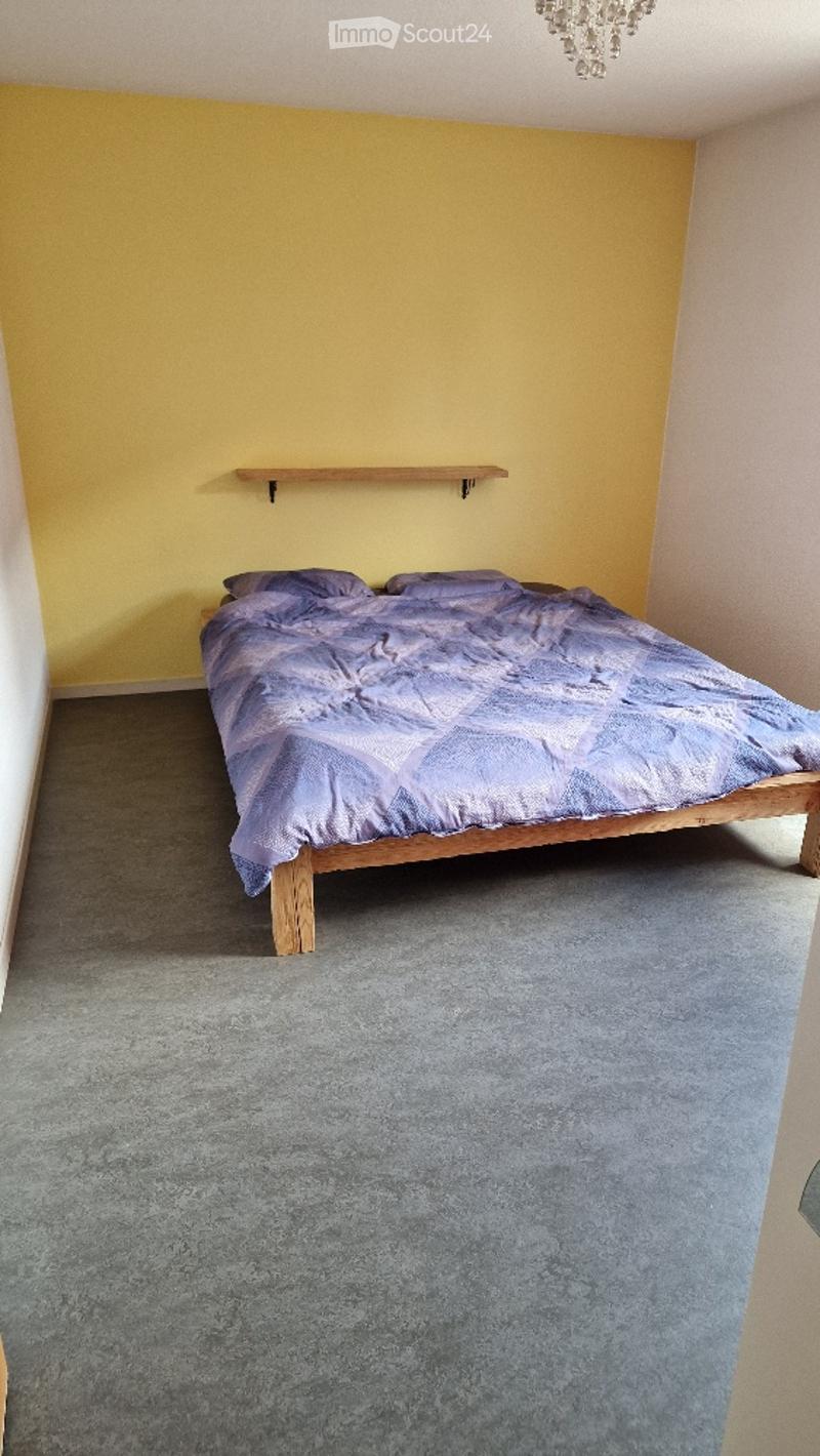 Bedroom with a wooden bed, purple bedding, a small wooden shelf on the wall, and a chandelier hanging from the ceiling.