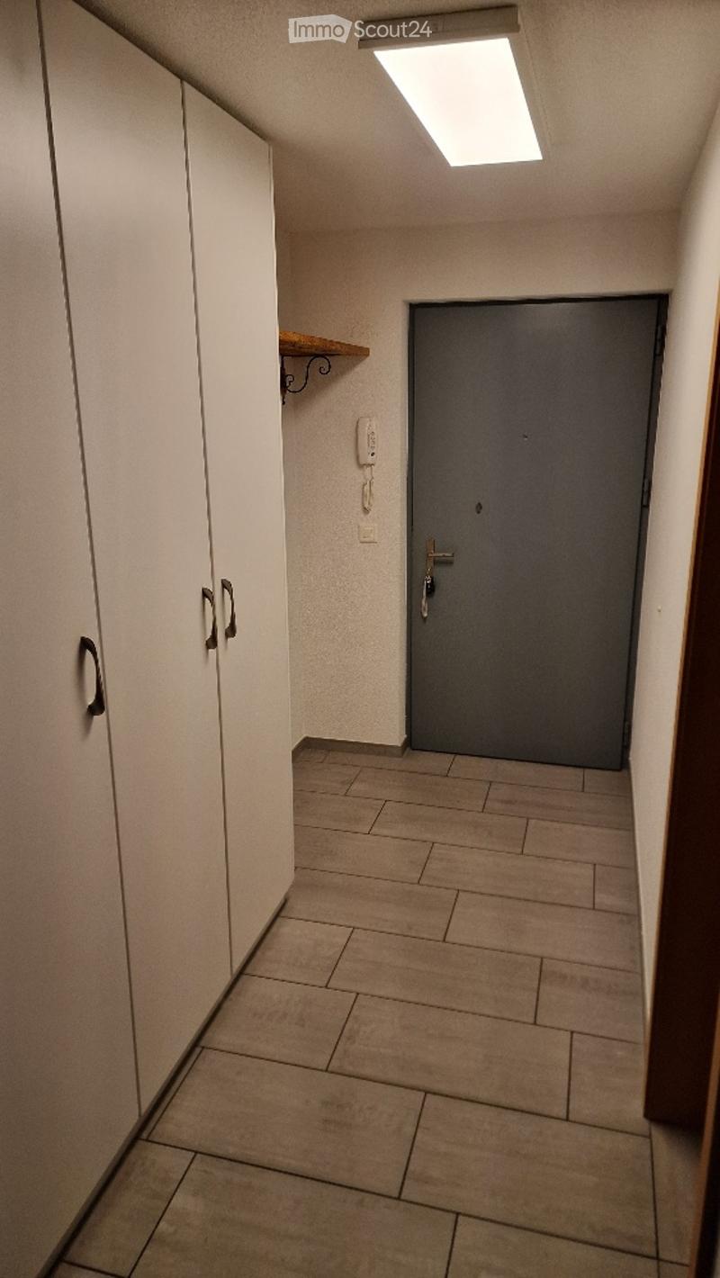 Empty hallway with tiled floor, white walls, a closed grey door, a small shelf, and a telephone mounted on the wall.