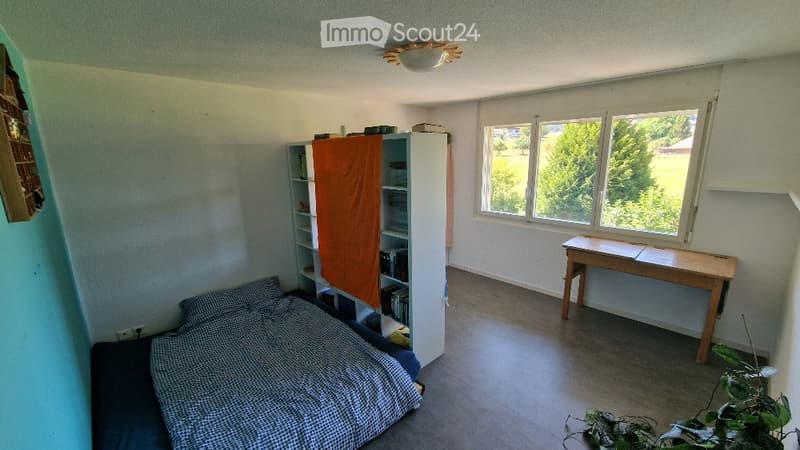 Bedroom with a bed, bookshelf, small table, and plant near the window. The room has large windows with a view of greenery outside.