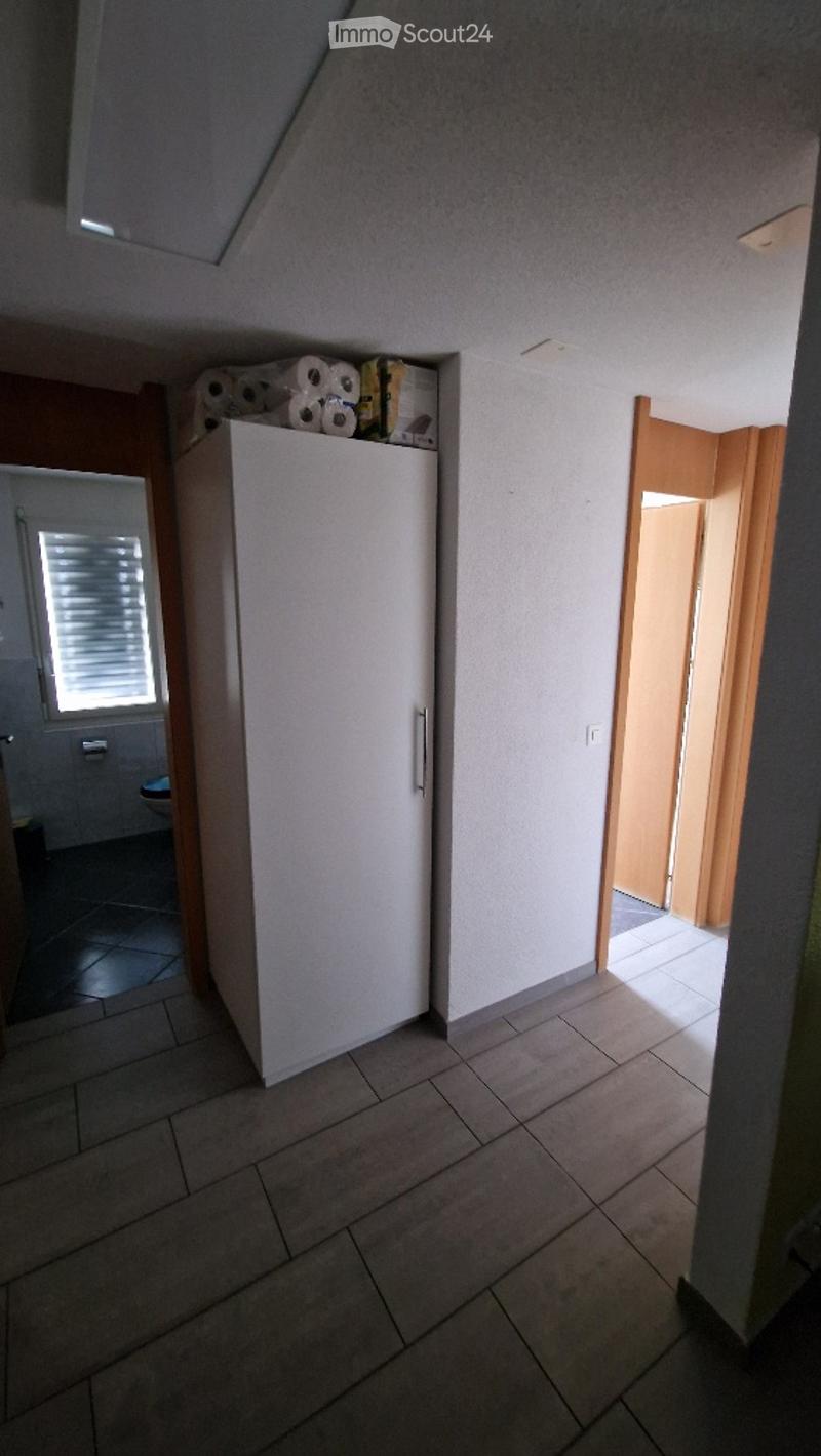 A small, tiled room with a white cupboard holding toilet paper and other items. The room has a window with blinds and a door leading to another area.