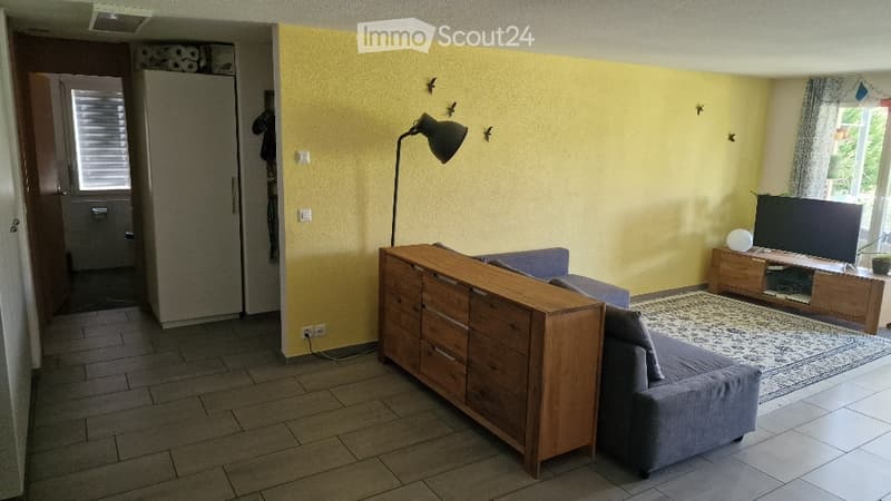Living room with a grey sofa, wooden cabinet, TV stand, floor lamp, and a small table. The walls are painted yellow, and there is a tiled floor.