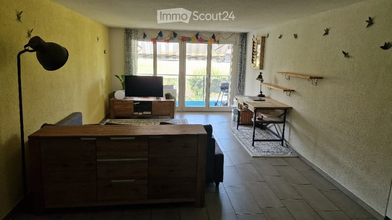 Bright living room with wooden furniture, large sliding glass doors leading to a patio, a sofa, coffee table, TV stand, and a small dining area with a table and chairs.