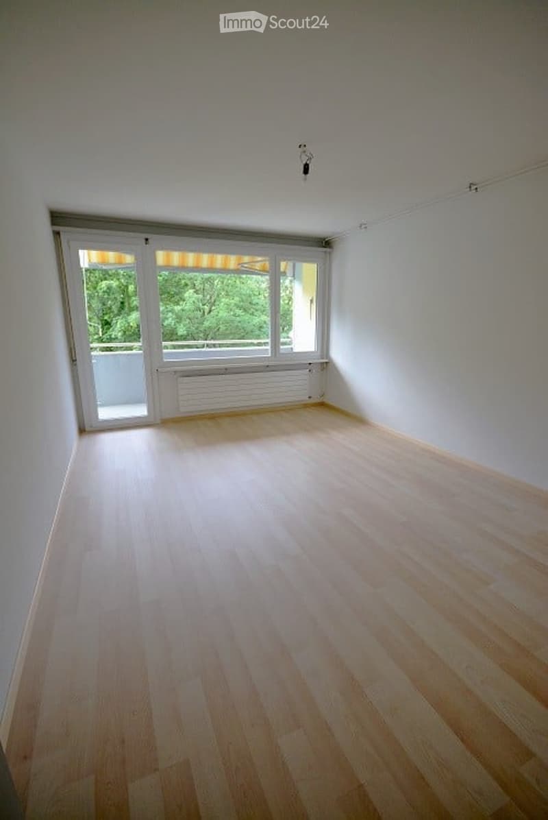 Empty room with light wooden flooring, white walls, large windows with a view of greenery, and a sliding door leading to a balcony.