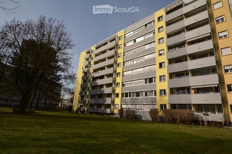 Multi-story residential building with yellow and gray facade, multiple windows, and balconies. Surrounded by green lawn and trees.