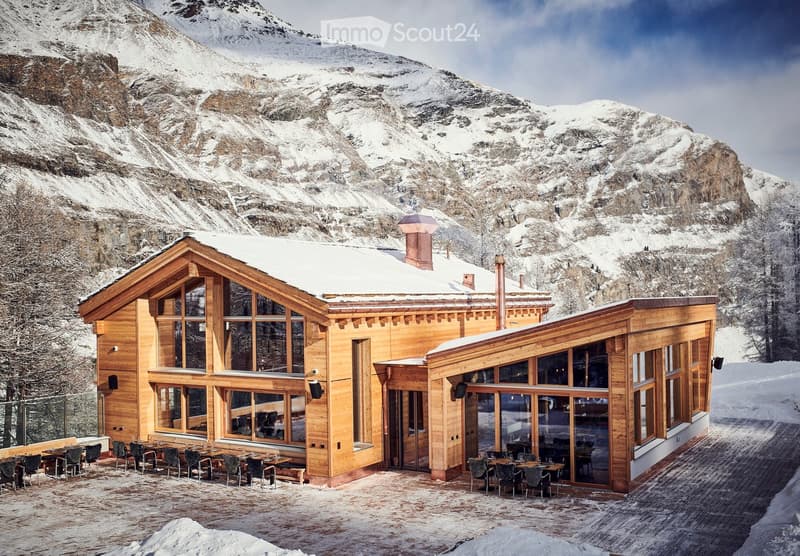 A large wooden building with snow-covered roof and large windows, situated in a snowy mountain landscape. The building has an outdoor seating area with tables and chairs.