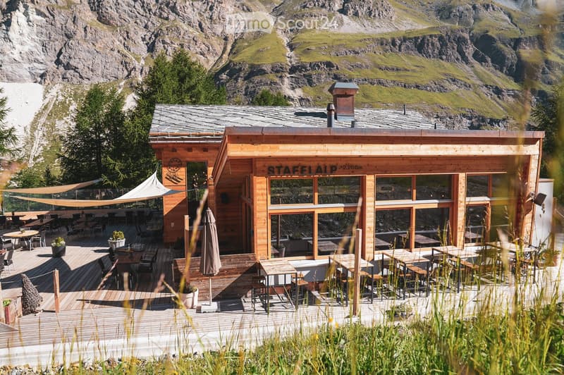 A wooden restaurant or cafe with large windows and a spacious outdoor seating area, surrounded by mountains and greenery.
