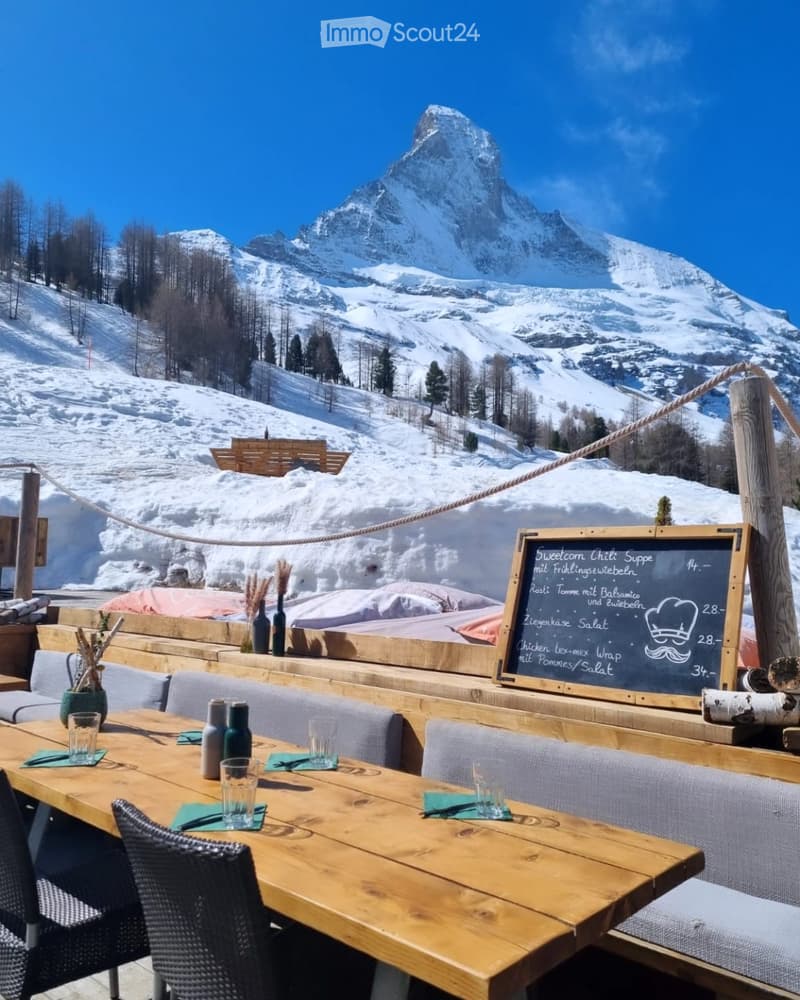 An outdoor dining area with wooden tables and chairs, set against a backdrop of snow-capped mountains and a forested landscape.