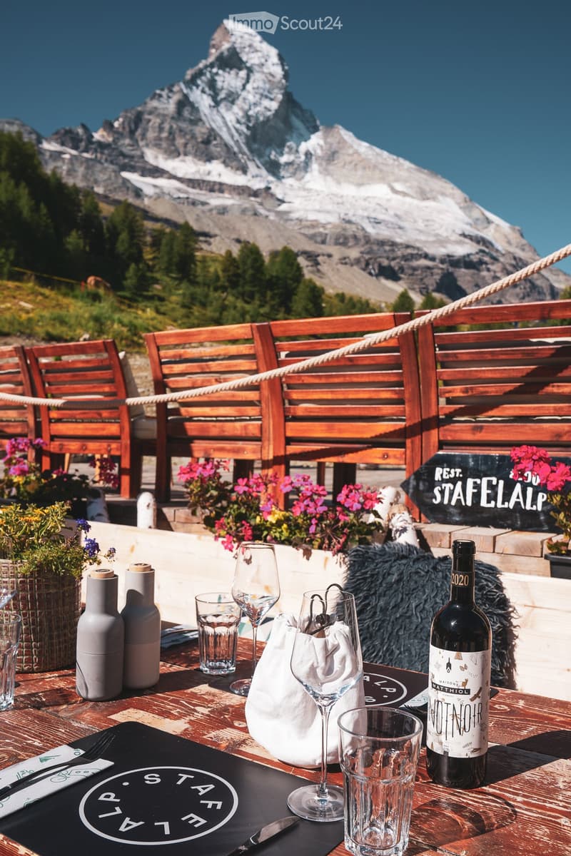 Outdoor seating area with wooden chairs and tables, overlooking a mountain range. Wine bottles, glasses, and a sign for "Rest. Stafelalp" are visible.
