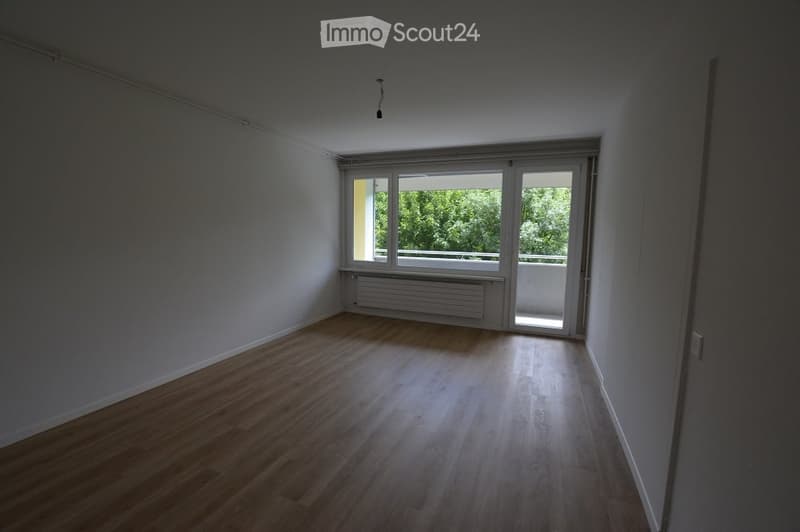 Empty room with wooden flooring, white walls, large window, and sliding glass door leading to a balcony.