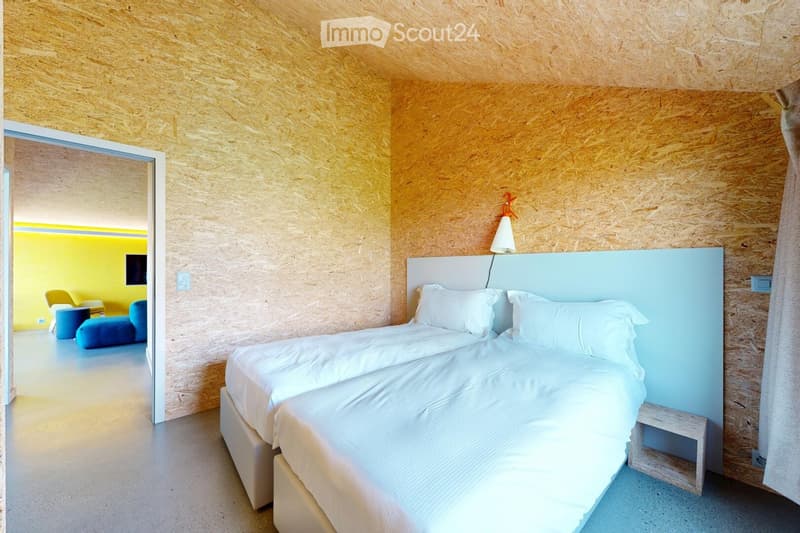 Modern bedroom with two single beds, white bedding, a light blue headboard, a small wooden nightstand, and a wall-mounted lamp. The walls are clad in light wood paneling, and the floor is light-colored concrete.
