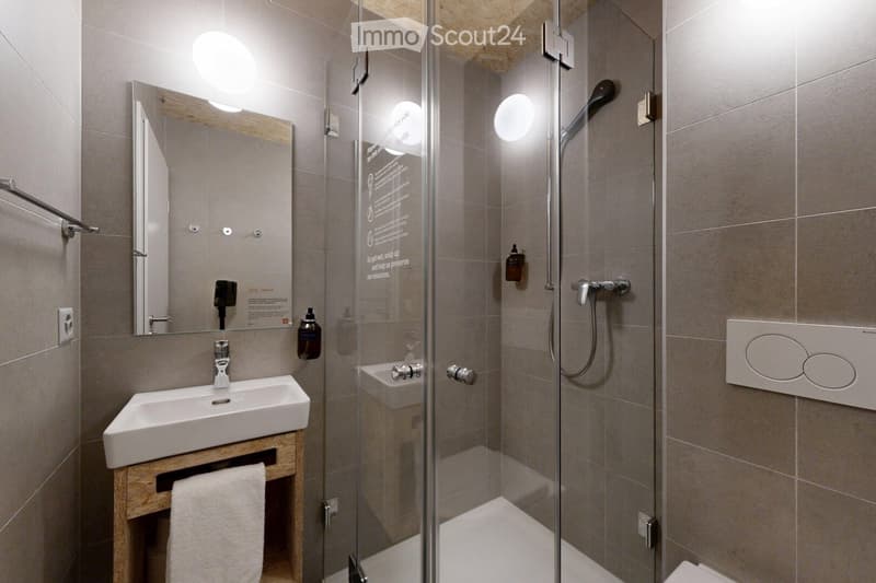 Modern bathroom with a glass-enclosed shower, a white sink on a wooden stand, a large mirror, and a towel hanging on the stand.