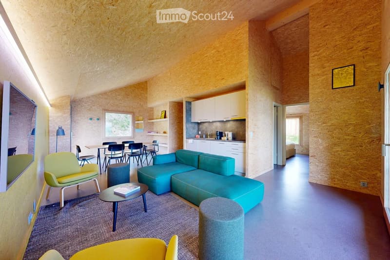 Modern living and dining area with a teal sectional sofa, coffee table, yellow armchair, dining table with chairs, and a small kitchen area with white cabinets and appliances.