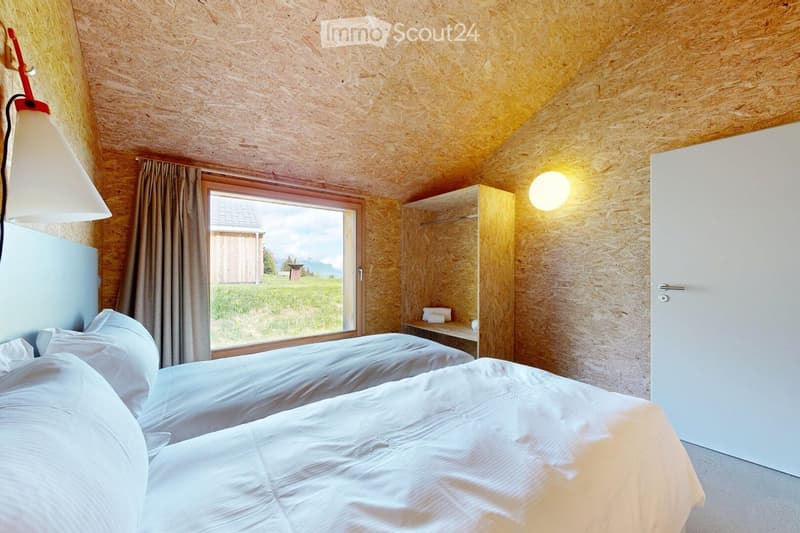 Cozy bedroom with wooden walls and ceiling, two single beds, a nightstand with a lamp, a wardrobe, and a large window with a view of a green field and mountains.