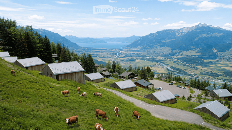 A scenic view of a rural area with cows grazing on a green hillside, surrounded by wooden barns and a winding road, with a vast valley and mountain range in the background.