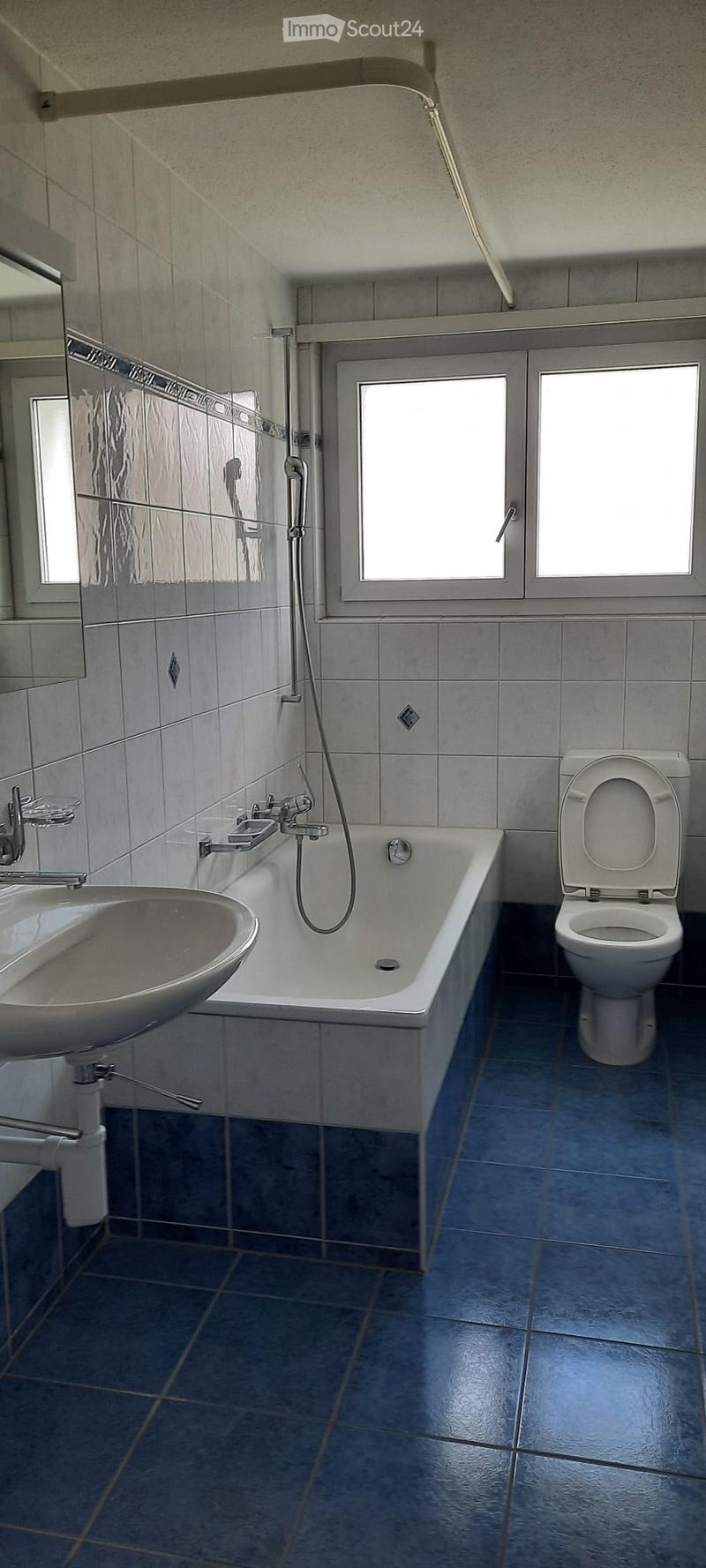 Modern bathroom with a bathtub, toilet, sink, and faucet. The room features grey tiled walls and blue tiled flooring, with a window providing natural light.