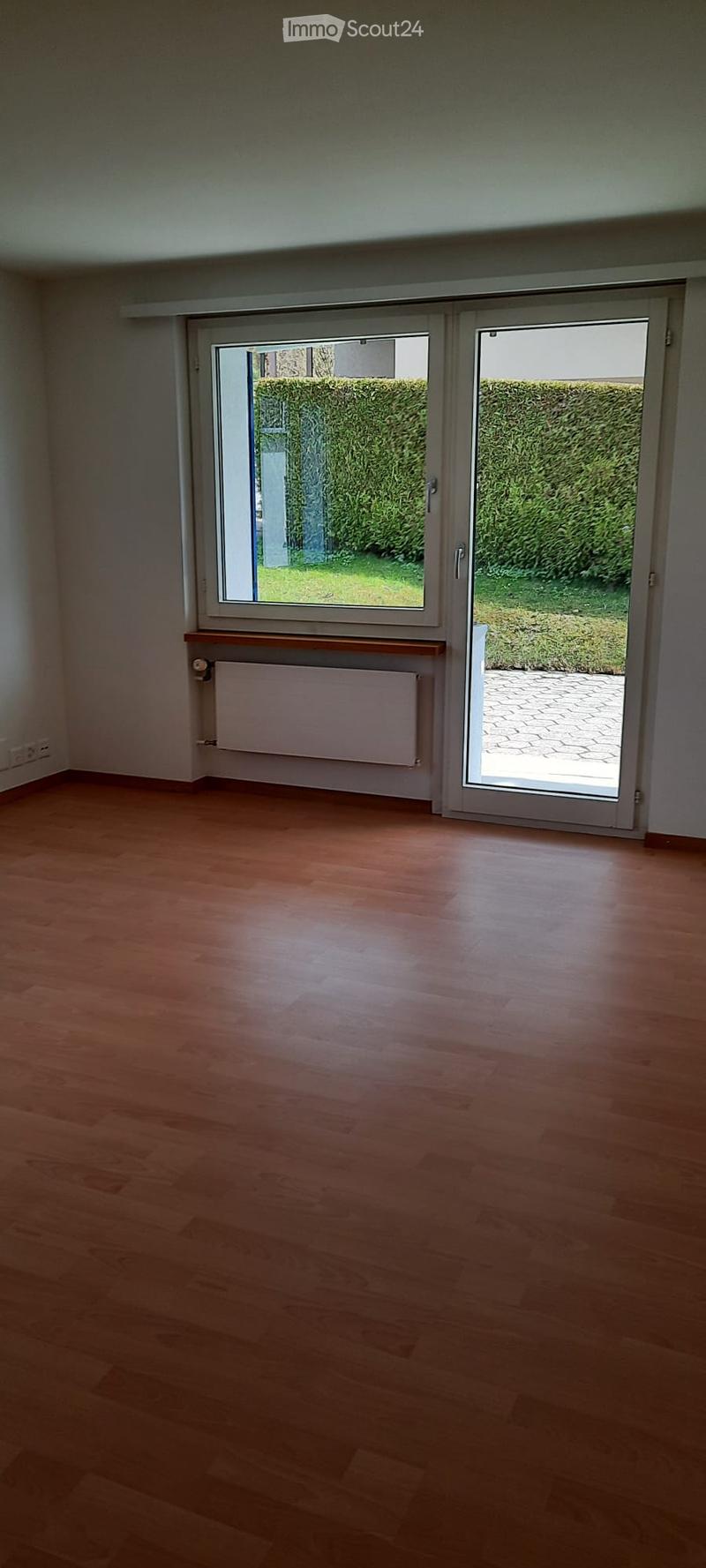 Empty room with wooden flooring, large sliding glass doors leading to a garden, radiator, and a small shelf above it.