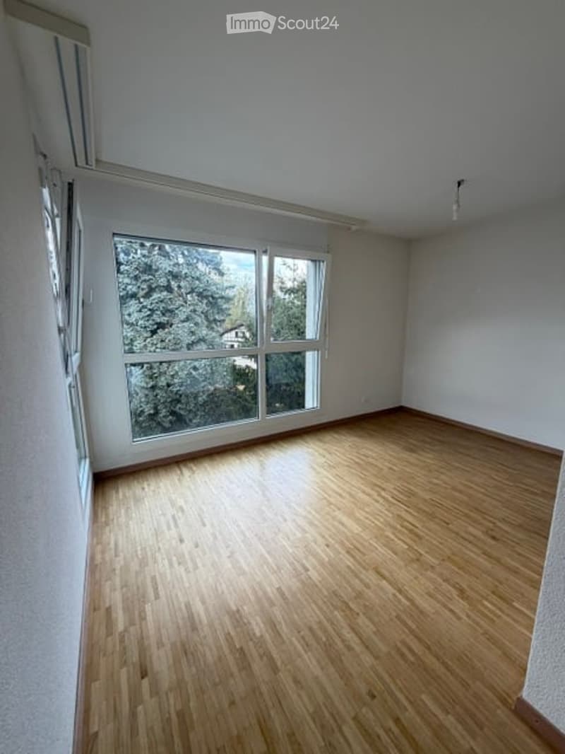 Empty room with wooden flooring and large windows showing a view of trees and a house outside.
