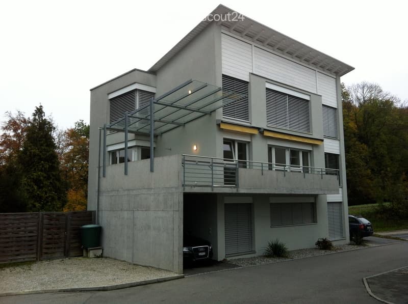 Modern two-story house with grey concrete facade, large windows, balcony, and integrated garage. Features a covered entrance and sleek design.