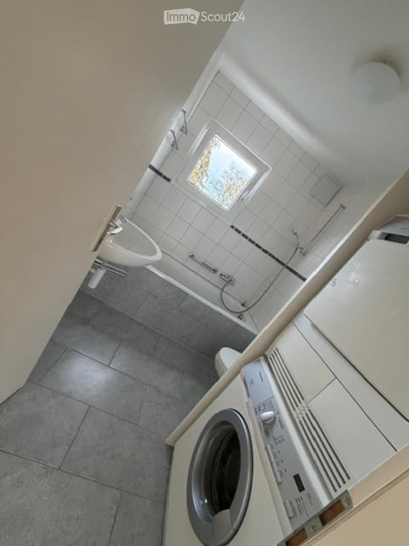 Bathroom with white tiled walls, grey tiled floor, a window, a sink, a toilet, a shower, and a washing machine.