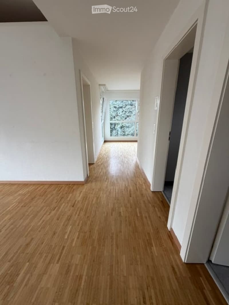 Empty hallway with wooden flooring, white walls, and a window at the end providing natural light.