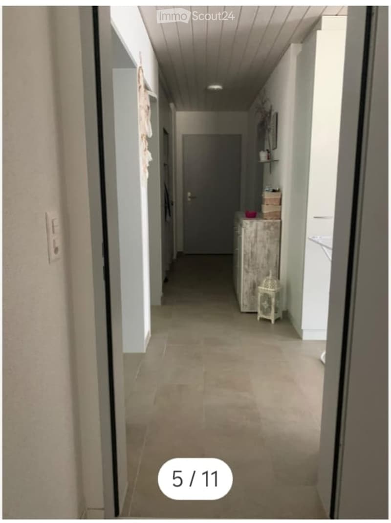 A long, narrow hallway with tiled flooring, white walls, and a closed door at the end. There is a small wooden cabinet with a decorative lantern and a few items on top, and a shelf with a few objects on the right side.