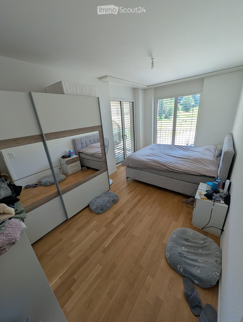 Bright bedroom with wooden flooring, large windows, a double bed with grey bedding, a wardrobe with a mirror, a nightstand, a bean bag chair, and a dog bed.