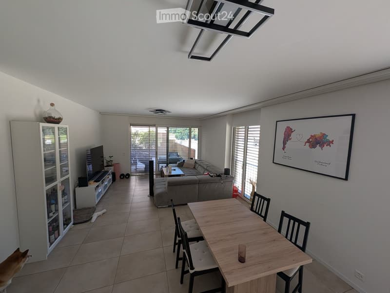 Modern living and dining area with a grey sectional sofa, coffee table, dining table with chairs, TV stand, and a cabinet. Large windows and sliding glass doors lead to an outdoor area.