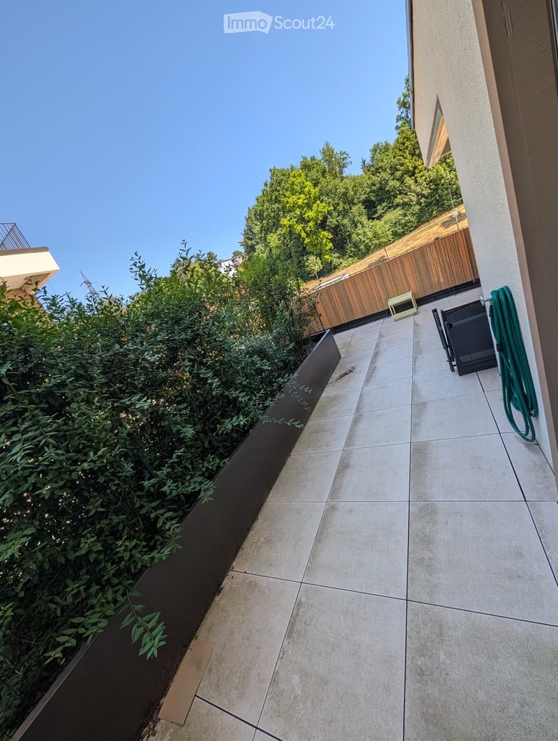 Modern outdoor patio with tiled flooring, green plants in a raised bed, wooden fence, and a green hose coiled against the wall.